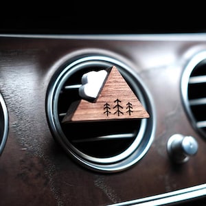 Puede incluir: Un difusor de ventilación de coche de madera con forma de montaña con tres árboles grabados. La montaña es marrón con una nube blanca. El difusor está colocado en una rejilla de ventilación del coche, con un salpicadero de madera oscura de fondo.