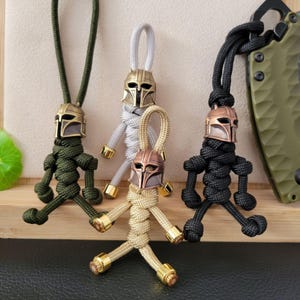 May include: Four handmade keychains featuring a Mandalorian helmet design. The keychains are made of braided cord in green, gray, gold, and black, with metallic gold accents. The helmets have a bronze finish.