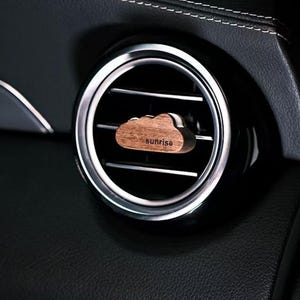 Könnte beinhalten: Ein holzfarbener, wolkenförmiger Auto-Lufterfrischer mit dem Aufdruck "sunrise". Der Lufterfrischer wird in die Lüftungsöffnung eines Autos eingesetzt. Die Lüftungsöffnung ist silber und schwarz, und der Innenraum des Autos ist schwarz.