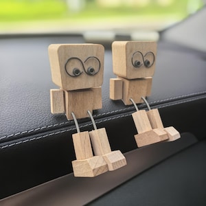 Puede incluir: Dos figuras de robot de madera con patas de alambre y ojos grandes y caprichosos. Los robots tienen un diseño simple y en bloques y están hechos de madera de color claro. Están sentados sobre una superficie oscura, posiblemente un salpicadero de coche, con un fondo verde borroso.