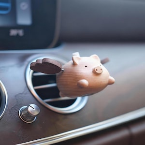 May include: A wooden pig car air vent clip with brown wooden wings. The pig has a small snout, ears, and a tail. The air vent clip is inserted into a car's air vent, which is surrounded by a silver trim.