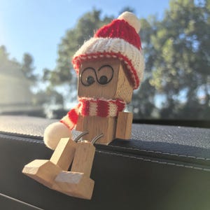 Puede incluir: Una figura de madera con cabeza cuadrada, que lleva un gorro y una bufanda de punto rojos y blancos. La figura tiene grandes ojos saltones y está sentada con las piernas extendidas. La figura es de madera clara y tiene un aspecto caprichoso y festivo.