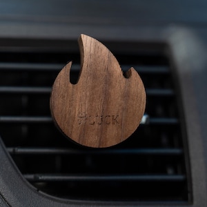 Puede incluir: Un clip de ventilación de coche de madera marrón con forma de llama. La palabra "LUCK" está grabada en el clip. El clip está sujeto a una rejilla de ventilación negra.