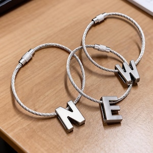 May include: Three silver keychains spelling the word "NEW". Each keychain features a silver metal letter attached to a silver cable loop. The keychains are displayed on a light brown wooden surface.