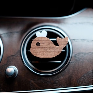 以下が含まれることがあります： 木製のクジラ型の車のエアベントクリップ。クジラは、頭に白いアクセントと濃い茶色の目を持つ明るい茶色の木材で作られています。クリップは、暗い木目調のダッシュボードを背景に、シルバーのトリムが付いた車のエアベントの上に配置されています。