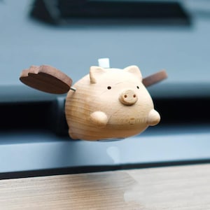 May include: A light brown wooden pig ornament with small, dark eyes and a detailed snout. The pig has small, brown wooden wings attached to its sides. The figurine appears to be floating.