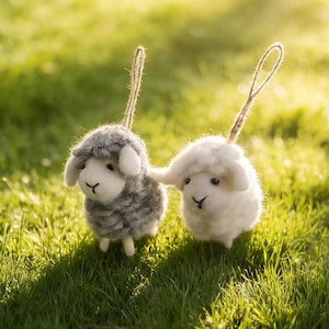 May include: Two small, fluffy sheep ornaments, one gray and one white, stand side-by-side on green grass. Each has a loop for hanging. The sheep have small black eyes and a stitched 'Y' shaped mouth.