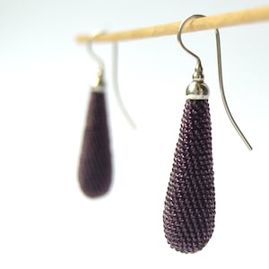 May include: A pair of dangle earrings with a teardrop-shaped bead made of dark purple beads. The earrings are silver and have a hook closure.