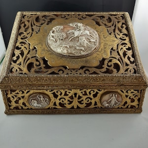 May include: A decorative box made of gold-toned metal with intricate carvings and a raised, oval-shaped panel featuring a white cameo of a hunting scene. The box has three smaller, circular panels with white cameos on the sides.