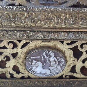 May include: A gold-toned decorative box with intricate floral designs and a cameo of a woman driving a chariot pulled by two horses.