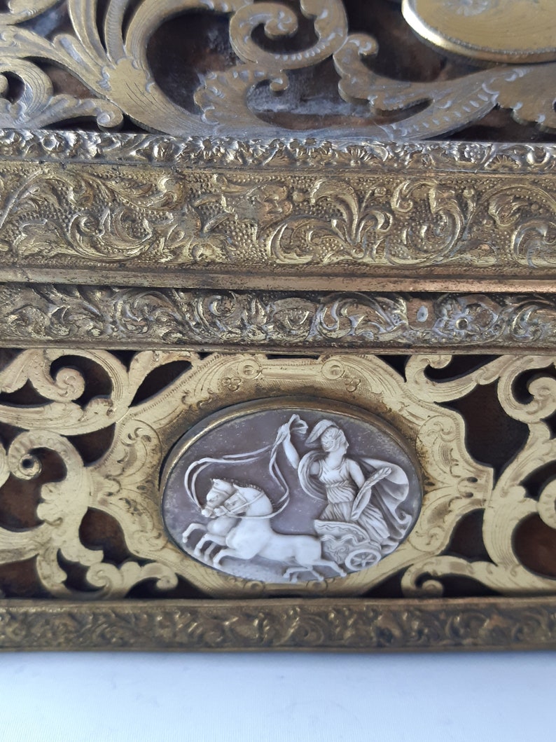 May include: A gold-toned decorative box with intricate floral designs and a cameo of a woman driving a chariot pulled by two horses.