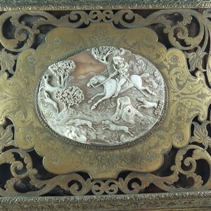 May include: A gold-toned metal box with an ornate, carved design. The box features a raised, oval-shaped panel with a carved scene of a rider on horseback chasing hounds through a forest.