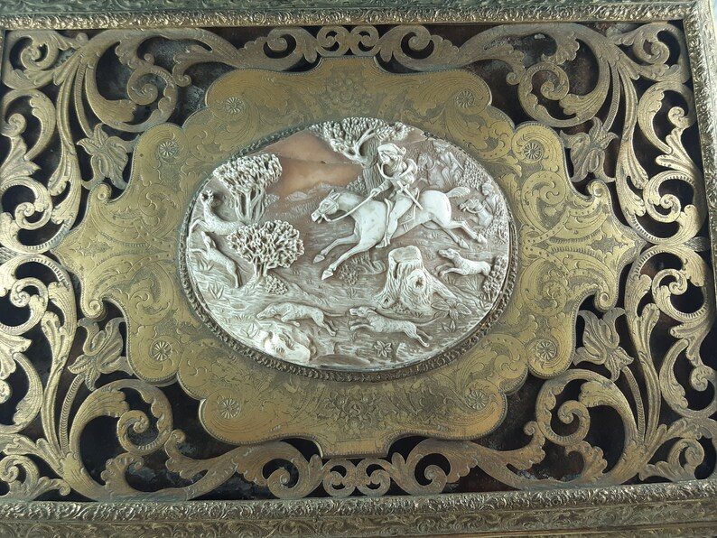 May include: A gold-toned metal box with an ornate, carved design. The box features a raised, oval-shaped panel with a carved scene of a rider on horseback chasing hounds through a forest.