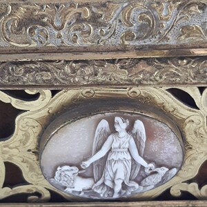 May include: An antique ornate gold-toned box with a cameo of a winged figure holding a lion and a bird. The box features intricate floral designs and a raised border.