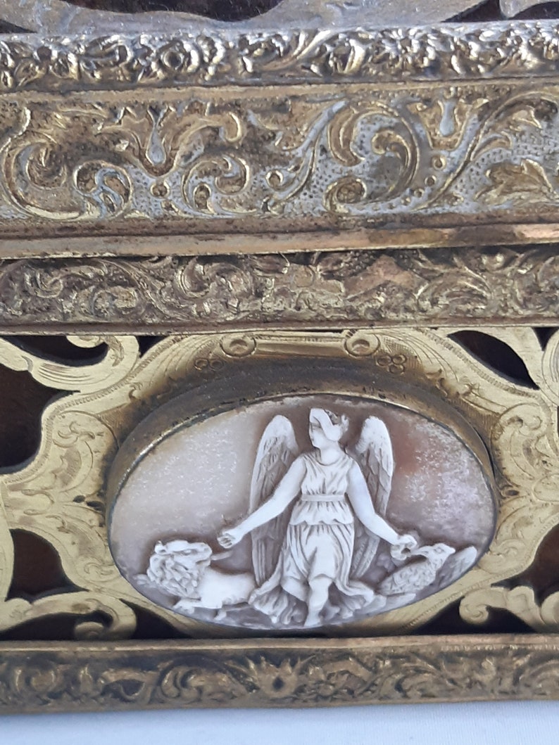 May include: An antique ornate gold-toned box with a cameo of a winged figure holding a lion and a bird. The box features intricate floral designs and a raised border.