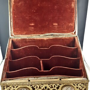 May include: An antique wooden box with a red velvet interior and intricate gold metalwork. The box has multiple compartments and is decorated with two oval medallions featuring raised figures.