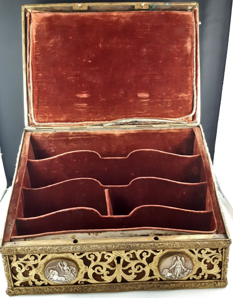May include: An antique wooden box with a red velvet interior and intricate gold metalwork. The box has multiple compartments and is decorated with two oval medallions featuring raised figures.