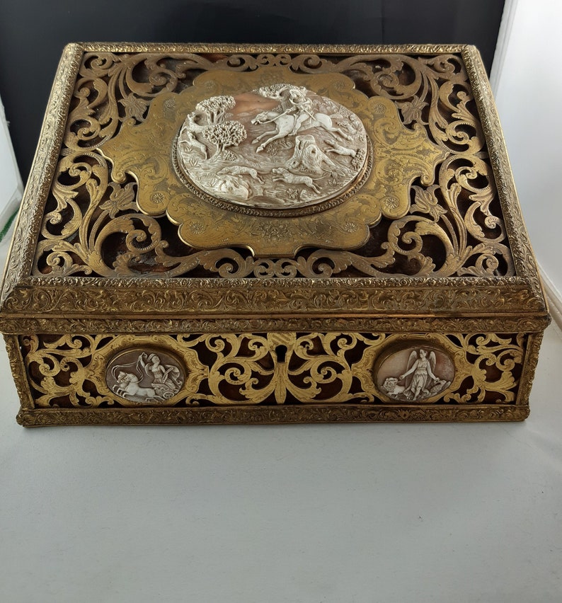 May include: A decorative box made of gold-toned metal with intricate carvings and a raised, oval-shaped panel featuring a white cameo of a hunting scene. The box has three smaller, circular panels with white cameos on the sides.