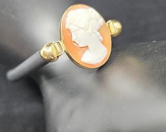 May include: An oval cameo ring featuring a white profile portrait of a person on an orange background, set in a gold-toned metal frame. The ring band is dark gray and connects to the central cameo setting with gold-toned metal accents.