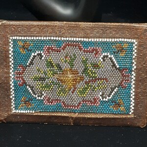 K&ouml;nnte beinhalten: Eine kleine, rechteckige, braune Lederbox mit einem Perlenmuster auf der Vorderseite. Das Muster zeigt ein florales Motiv in den Farben Blau, Gr&uuml;n, Wei&szlig; und Rosa.