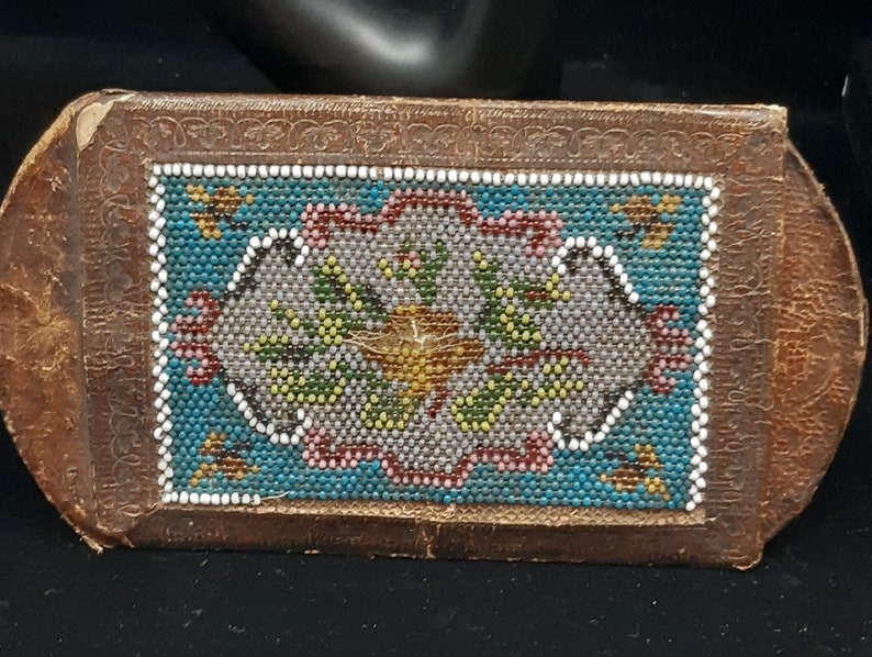 K&ouml;nnte beinhalten: Eine kleine, rechteckige, braune Lederbox mit einem Perlenmuster auf der Vorderseite. Das Muster zeigt ein florales Motiv in den Farben Blau, Gr&uuml;n, Wei&szlig; und Rosa.