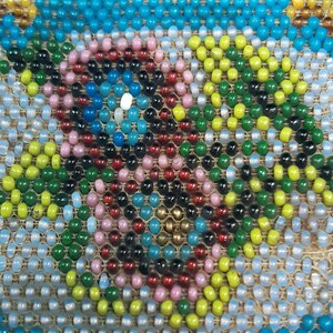 K&ouml;nnte beinhalten: Nahaufnahme eines Perlenmusters mit einem kreisf&ouml;rmigen Muster aus rosa, roten, schwarzen, blauen, gr&uuml;nen und gelben Perlen. Die Perlen sind auf einen beigefarbenen Stoffgrund gen&auml;ht.