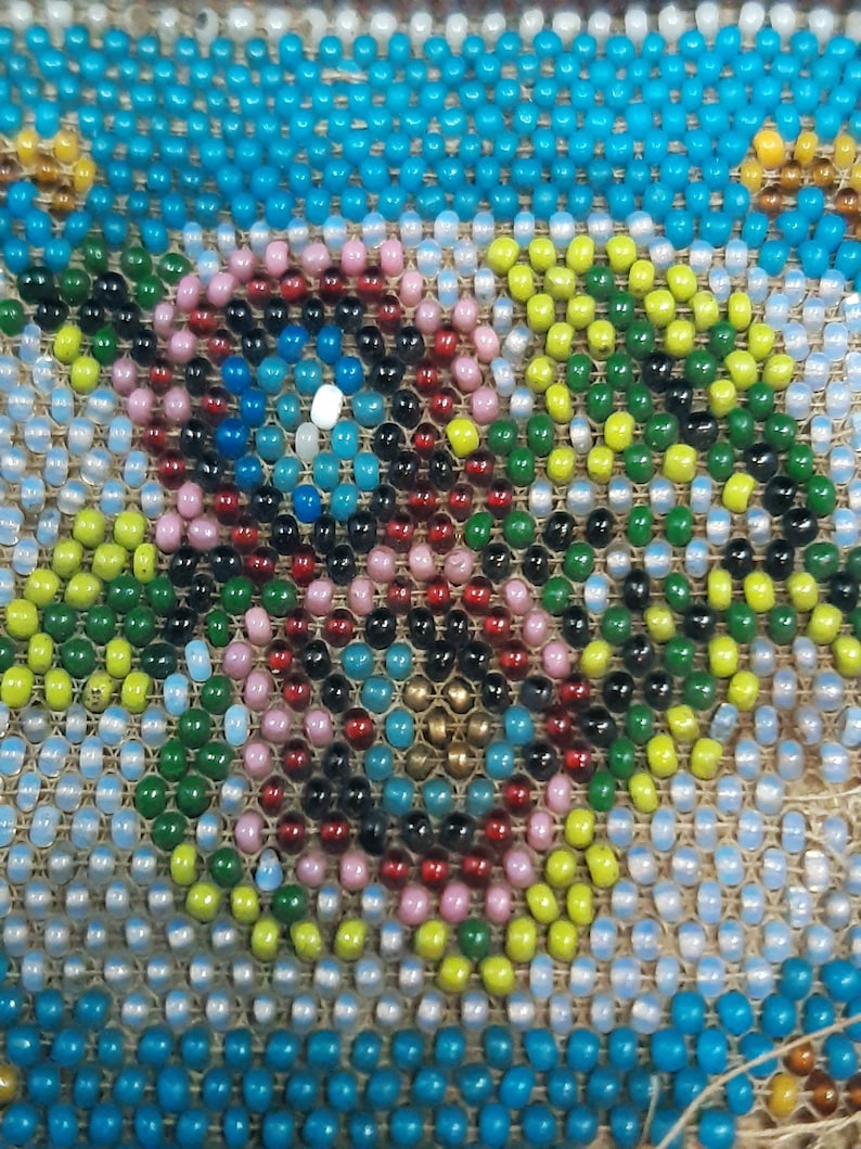 K&ouml;nnte beinhalten: Nahaufnahme eines Perlenmusters mit einem kreisf&ouml;rmigen Muster aus rosa, roten, schwarzen, blauen, gr&uuml;nen und gelben Perlen. Die Perlen sind auf einen beigefarbenen Stoffgrund gen&auml;ht.