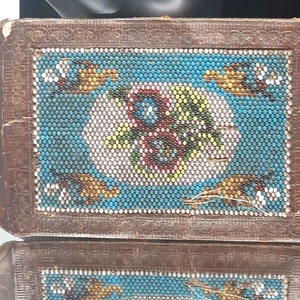 K&ouml;nnte beinhalten: Eine kleine, rechteckige, braune Lederbox mit einem Perlenmuster auf der Oberseite. Das Perlenmuster zeigt einen blauen Hintergrund mit einem floralen Muster in Wei&szlig;, Gelb, Gr&uuml;n und Rot.