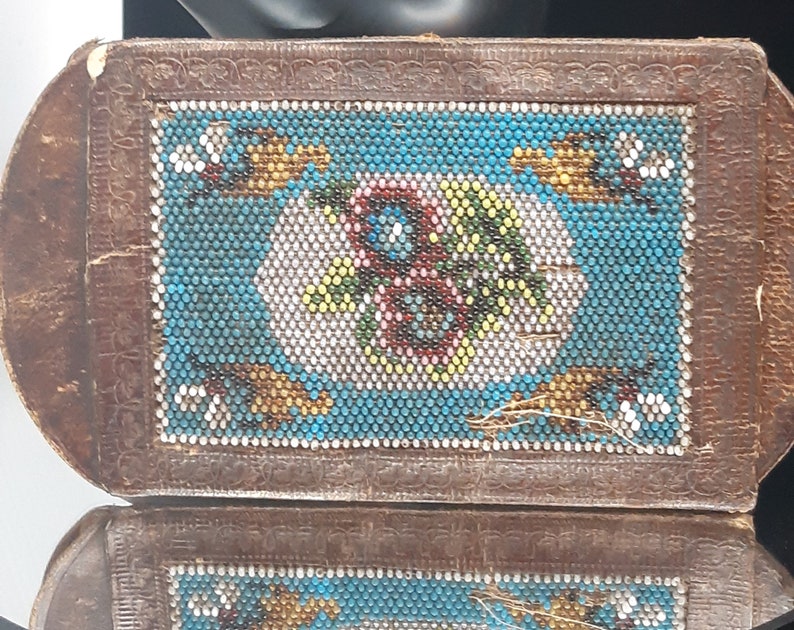 K&ouml;nnte beinhalten: Eine kleine, rechteckige, braune Lederbox mit einem Perlenmuster auf der Oberseite. Das Perlenmuster zeigt einen blauen Hintergrund mit einem floralen Muster in Wei&szlig;, Gelb, Gr&uuml;n und Rot.