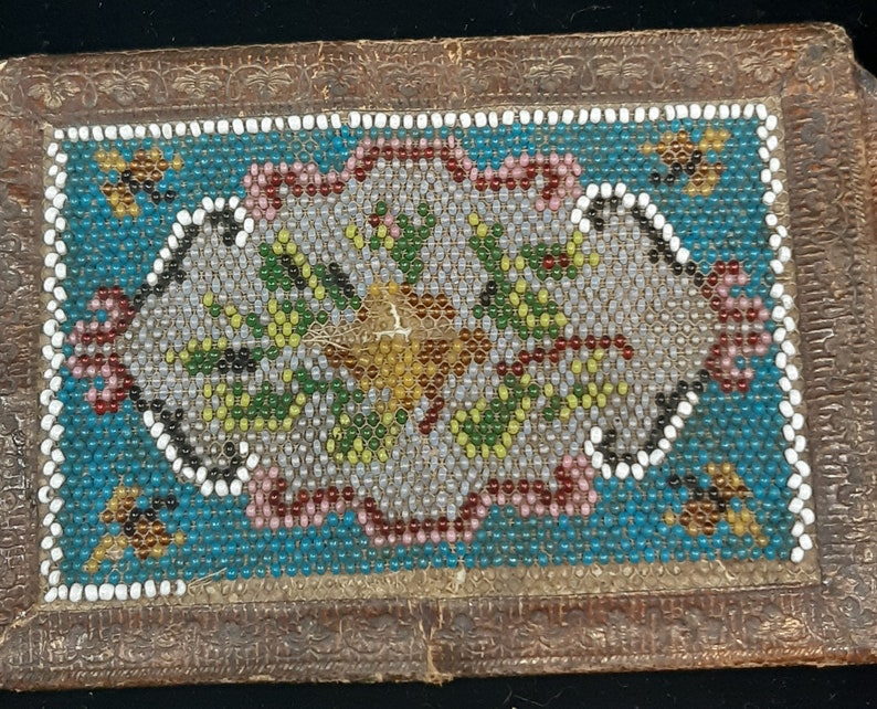 K&ouml;nnte beinhalten: Eine kleine, rechteckige Schachtel mit einem Perlenmuster. Die Schachtel ist aus braunem Leder und hat ein florales Muster in der Mitte. Die Perlen sind in verschiedenen Farben, darunter Wei&szlig;, Blau, Gr&uuml;n und Rot.