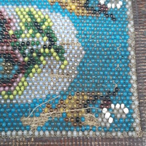 K&ouml;nnte beinhalten: Nahaufnahme eines Perlenmusters mit verschiedenen Farben, darunter Blau, Wei&szlig;, Gr&uuml;n und Braun. Die Perlen sind in einem geometrischen Muster angeordnet und auf einen Stoffuntergrund gen&auml;ht.