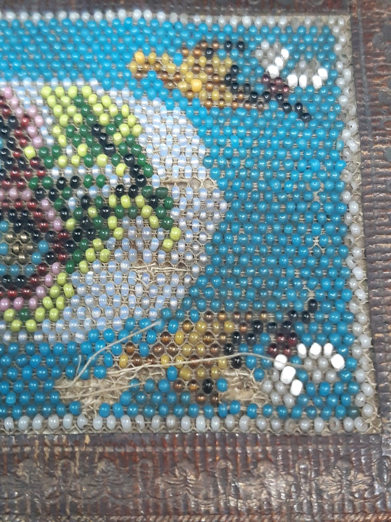 K&ouml;nnte beinhalten: Nahaufnahme eines Perlenmusters mit verschiedenen Farben, darunter Blau, Wei&szlig;, Gr&uuml;n und Braun. Die Perlen sind in einem geometrischen Muster angeordnet und auf einen Stoffuntergrund gen&auml;ht.