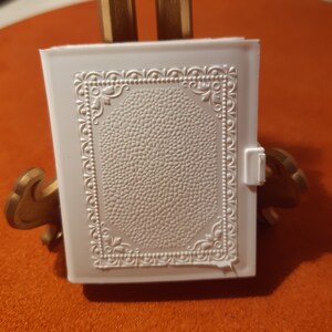 Puede incluir: Una caja de plástico blanca con un diseño en relieve texturizado y un borde decorativo. La caja tiene una tapa con bisagras y un pequeño pestillo.