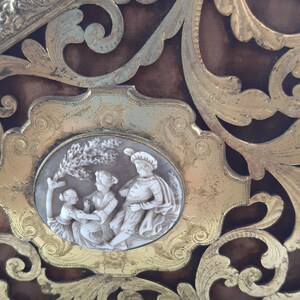 May include: A gold-toned decorative frame with an ornate design. The frame features a cameo with a white background and three figures in relief. The figures are dressed in white and are depicted in a playful scene.