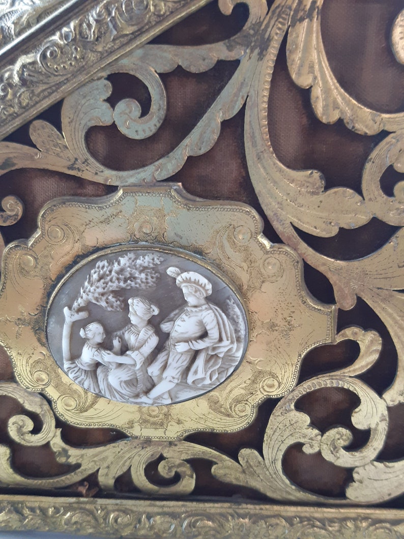 May include: A gold-toned decorative frame with an ornate design. The frame features a cameo with a white background and three figures in relief. The figures are dressed in white and are depicted in a playful scene.