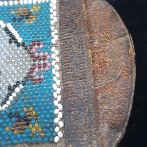 K&ouml;nnte beinhalten: Eine kleine, blaue und wei&szlig;e Perlenb&ouml;rse mit einer braunen Lederklappe. Die Klappe hat ein dekoratives Blumenmuster.