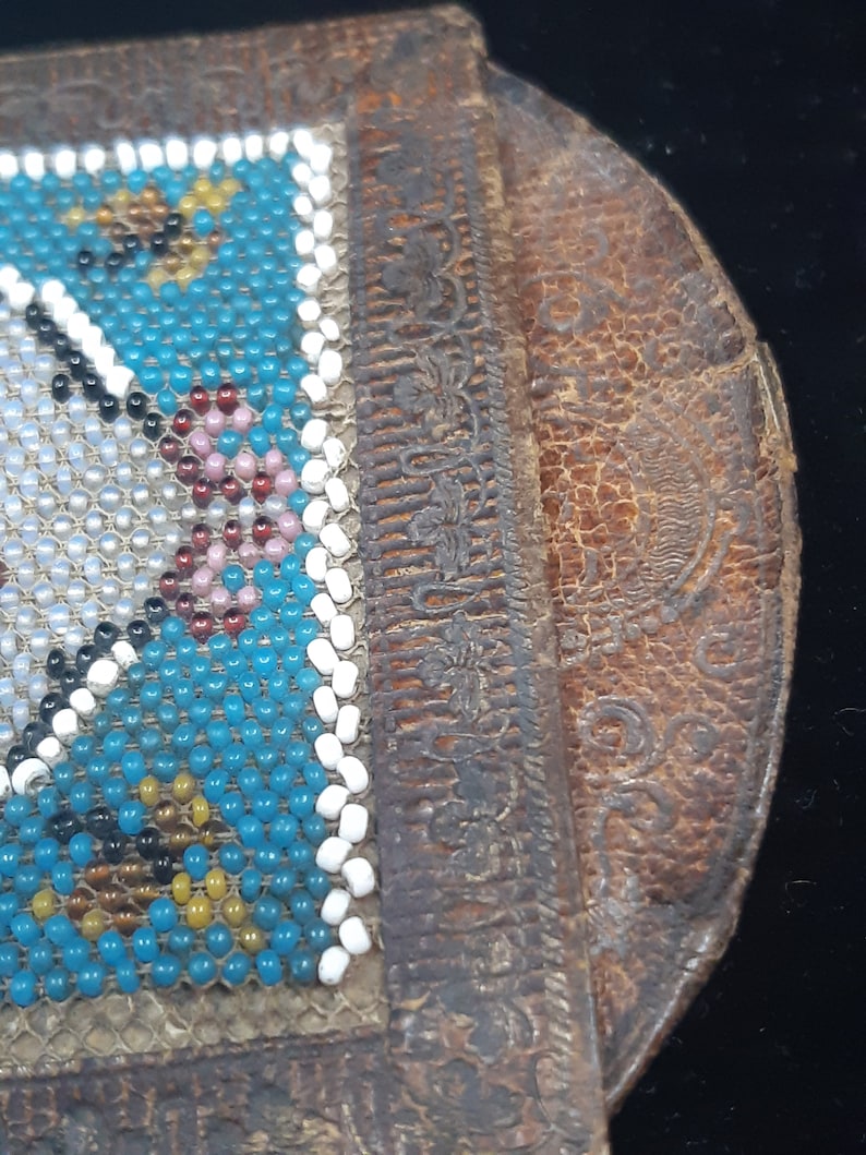 K&ouml;nnte beinhalten: Eine kleine, blaue und wei&szlig;e Perlenb&ouml;rse mit einer braunen Lederklappe. Die Klappe hat ein dekoratives Blumenmuster.