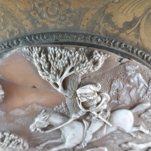 May include: A white cameo featuring a detailed carving of a person riding a horse through a woodland setting. The cameo is set in a gold-toned metal frame with an ornate design.