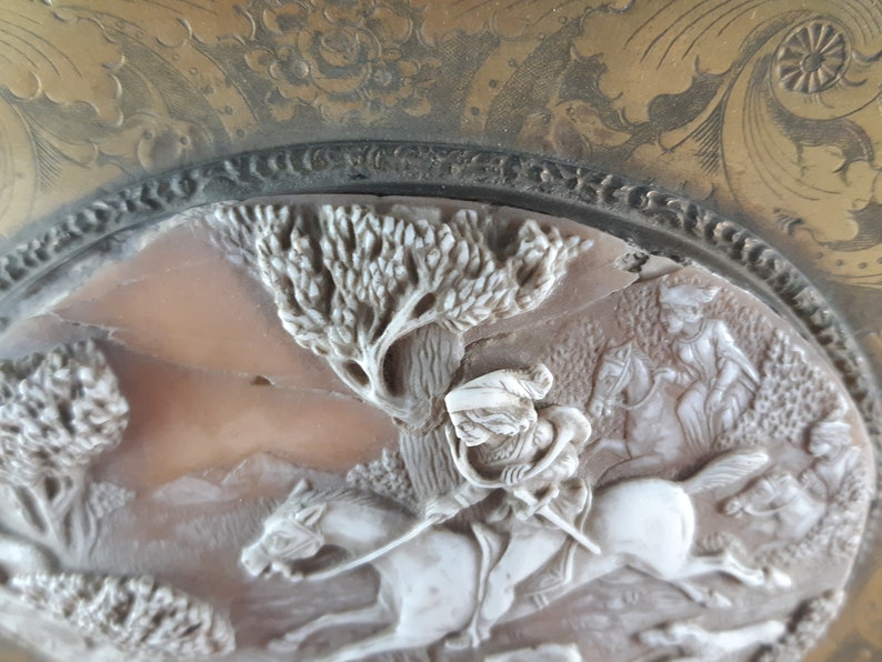 May include: A white cameo featuring a detailed carving of a person riding a horse through a woodland setting. The cameo is set in a gold-toned metal frame with an ornate design.