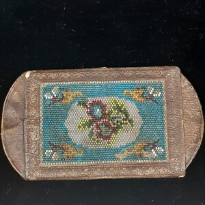 K&ouml;nnte beinhalten: Eine kleine, rechteckige, braune Lederbox mit einem Perlenmuster auf der Vorderseite. Das Muster zeigt ein florales Motiv in den Farben Blau, Gr&uuml;n, Gelb und Rot.