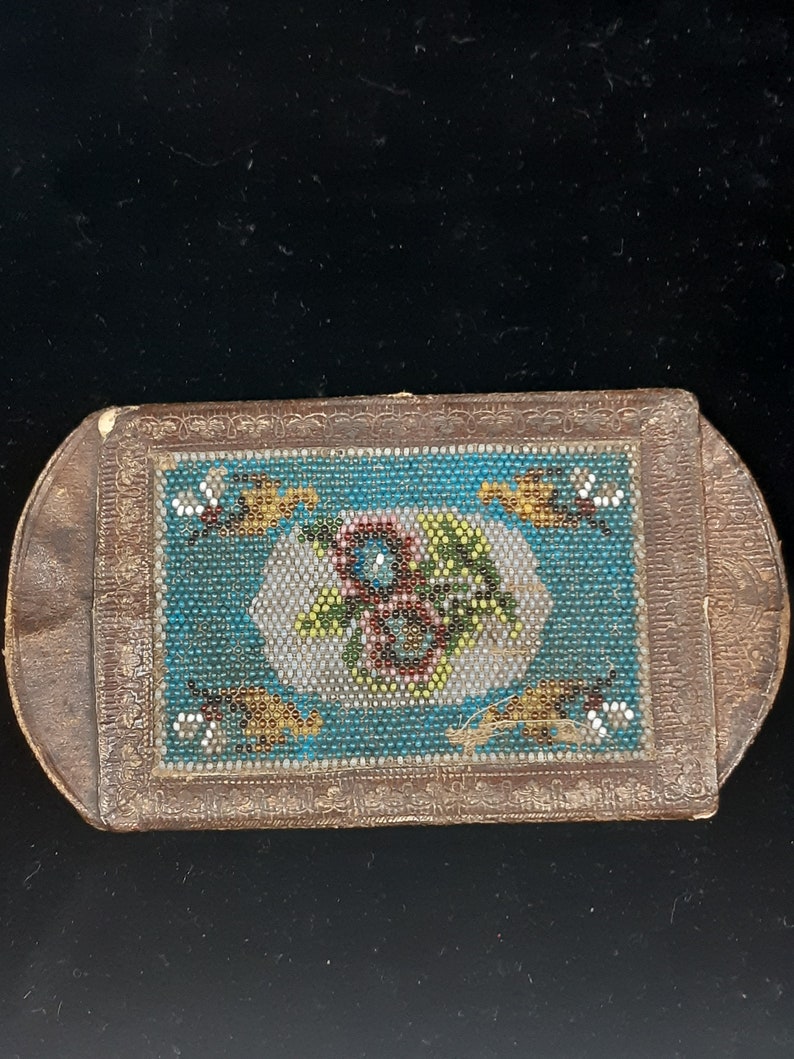 K&ouml;nnte beinhalten: Eine kleine, rechteckige, braune Lederbox mit einem Perlenmuster auf der Vorderseite. Das Muster zeigt ein florales Motiv in den Farben Blau, Gr&uuml;n, Gelb und Rot.