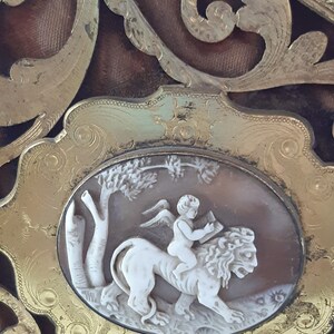 May include: An oval frame with intricate gold scrollwork surrounds a cameo depicting a winged cherub riding a lion. The cameo is carved in white and beige tones.