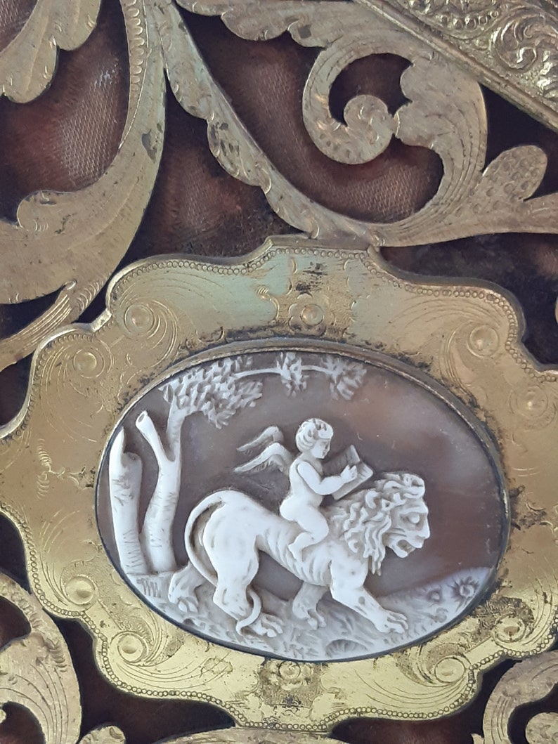 May include: An oval frame with intricate gold scrollwork surrounds a cameo depicting a winged cherub riding a lion. The cameo is carved in white and beige tones.