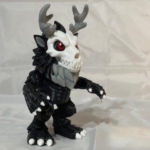May include: A small figurine of a creature with a white skull-like head, grey antlers, and red eyes. The body is black with textured fur-like details, white claws on the hands and feet, and a white chest plate. The figure stands in a semi-crouched pose.