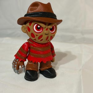May include: A stylized vinyl figure of a character wearing a brown fedora, a red and brown striped sweater, and brown trousers. The figure has a scarred, textured face with red eyes and a right hand featuring metallic-colored clawed fingers.