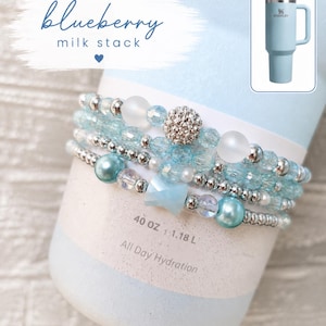 May include: A stack of light blue beaded bracelets with silver accents and a sparkling bead, displayed on a light blue tumbler. The tumbler has a handle and a straw. The text "blueberry milk stack" is visible.