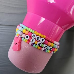 May include: Pink tumbler with a pink and white glitter finish. The tumbler is decorated with a multi-colored beaded bracelet that spells out "HEY PEEPS". A pink Peeps bunny charm is attached to the bracelet.