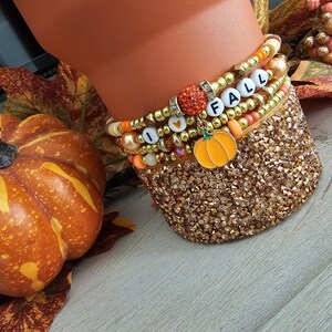 May include: A set of four stacked beaded bracelets featuring metallic gold, orange, and cream-colored beads. The bracelets include white letter beads spelling I HEART FALL, a sparkling orange sphere bead, and a dangling orange pumpkin charm.