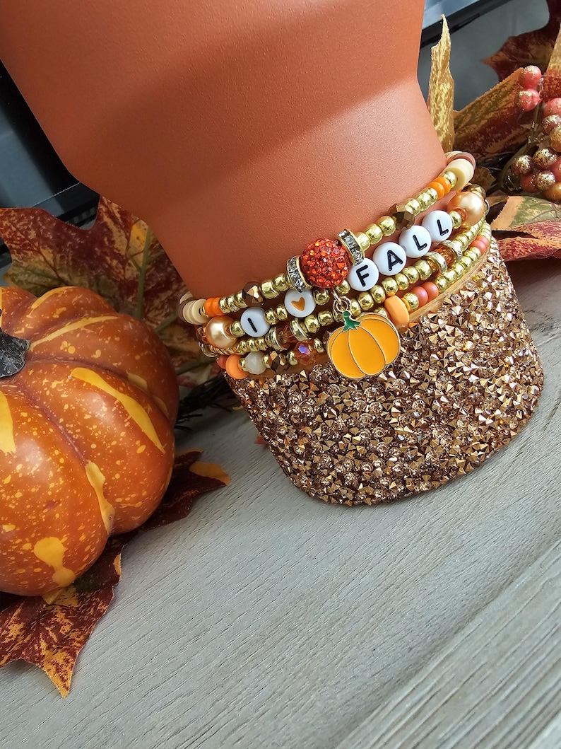 May include: A set of four stacked beaded bracelets featuring metallic gold, orange, and cream-colored beads. The bracelets include white letter beads spelling I HEART FALL, a sparkling orange sphere bead, and a dangling orange pumpkin charm.