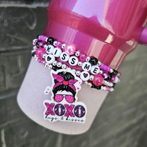 Könnte beinhalten: Rosa-schwarzes Perlenarmband mit den Worten "Kiss Me" und einem rosa-schwarzen Cartoon-Mädchen-Anhänger mit den Worten "XOXO hugs & kisses".